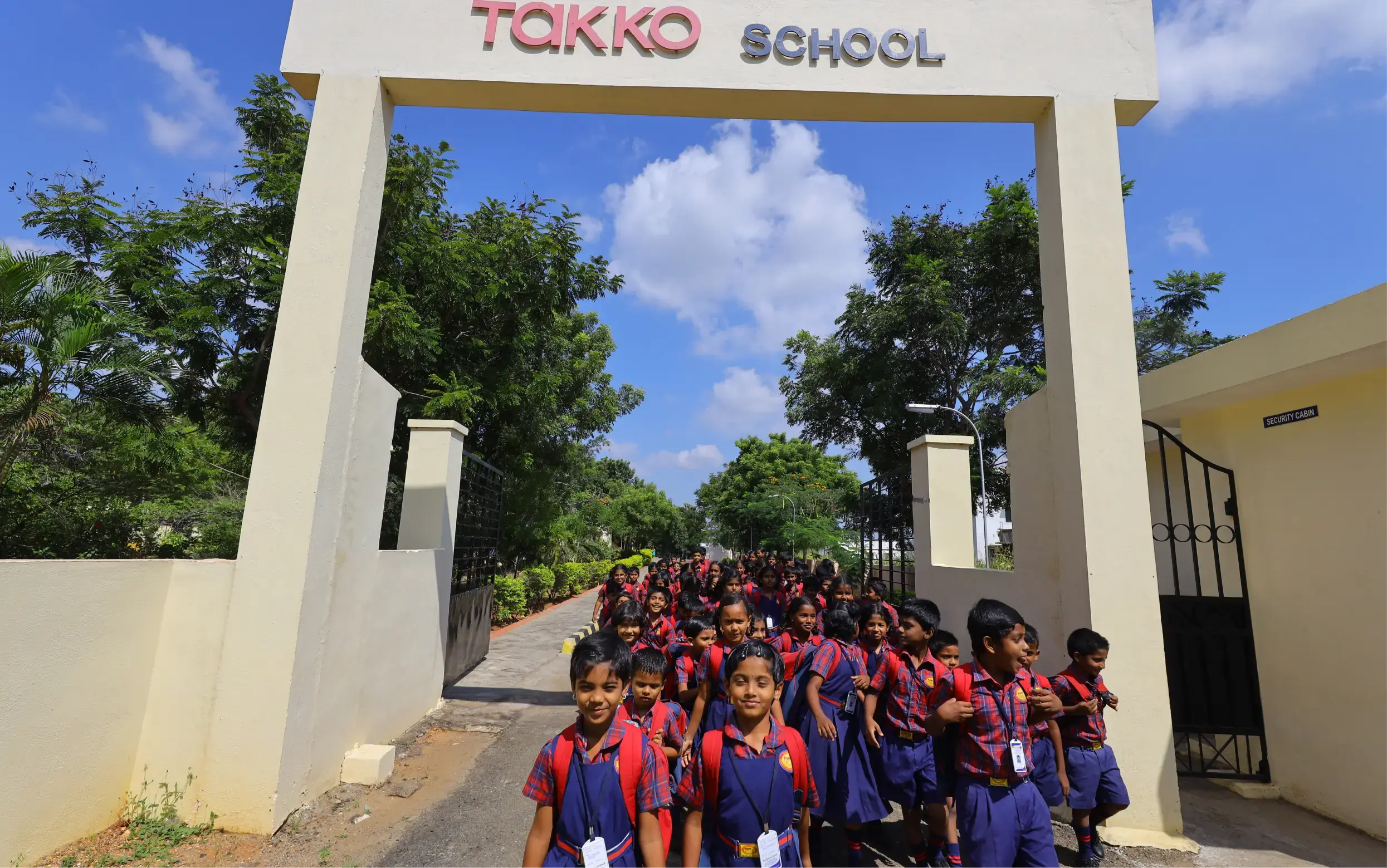 Un grand groupe de jeunes étudiants en uniforme sort d'une porte d'école étiquetée TAKKO SCHOOL par une journée ensoleillée, avec des arbres et un ciel bleu en arrière-plan.