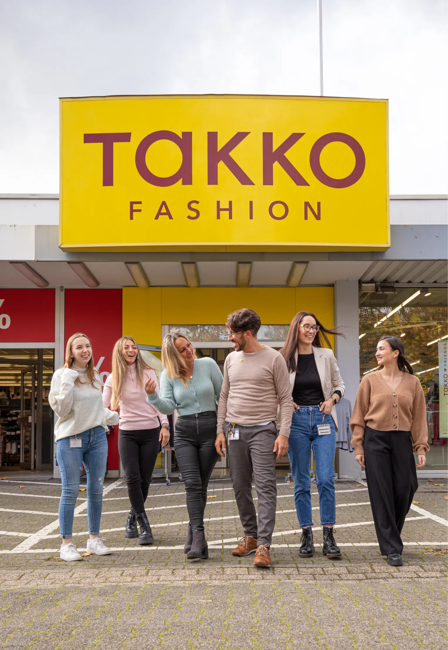 Sechs lächelnde Menschen gehen gemeinsam vor einem Takko Fashion-Geschäft mit einem großen gelb-roten Takko Fashion-Schild über dem Eingang.