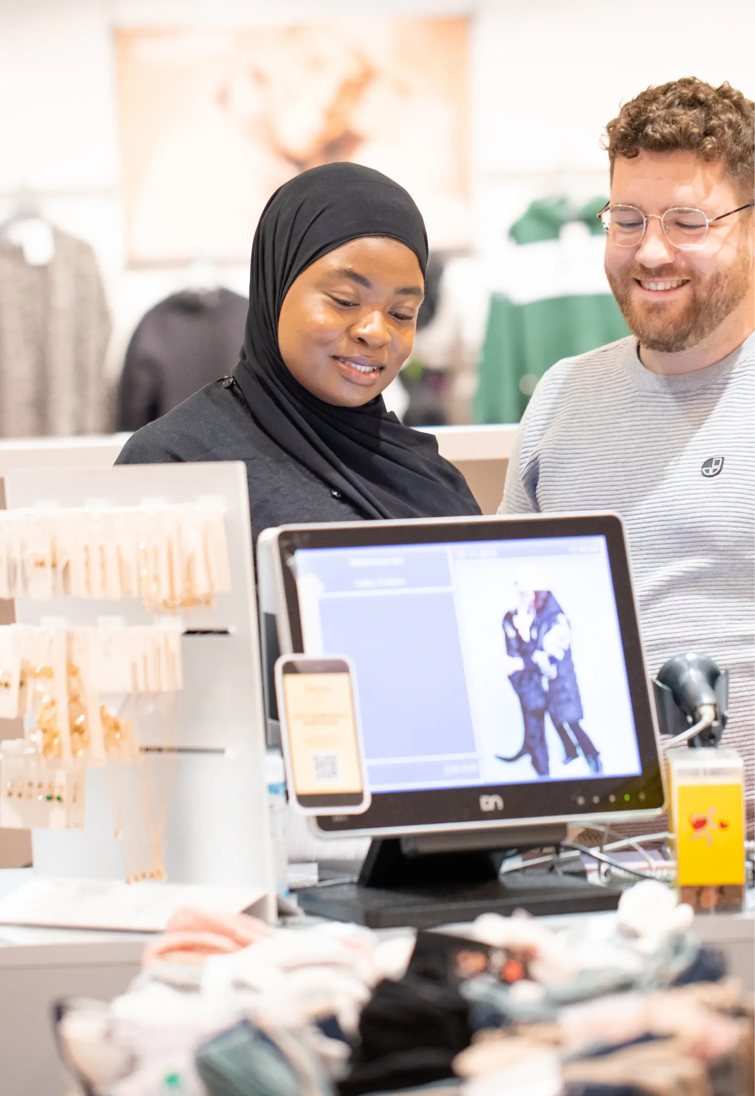 Eine Frau mit schwarzem Hijab und ein Mann mit Brille lächeln, während sie in einem Geschäft auf einen Computerbildschirm schauen, umgeben von Schmuckauslagen und Accessoires.