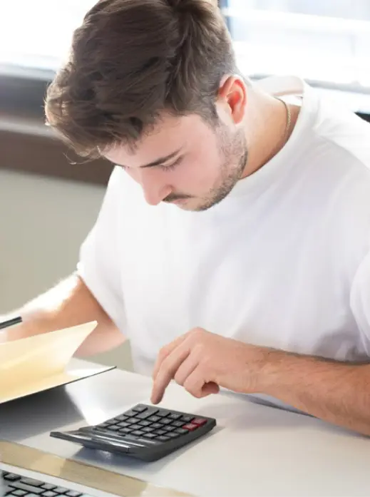 Ein Mann in einem weißen Hemd sitzt an einem Schreibtisch, blickt nach unten und drückt Tasten auf einem Taschenrechner, vor ihm liegt ein geöffneter Ordner.