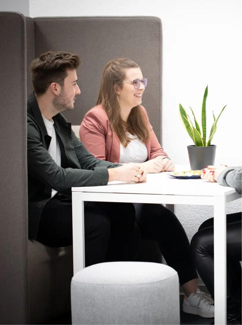 Zwei Personen sitzen nebeneinander an einem weißen Tisch in einer modernen, hellen Bürokabine und unterhalten sich mit einer dritten Person, die ihnen den Rücken zukehrt. Auf dem Tisch steht eine grüne Topfpflanze.