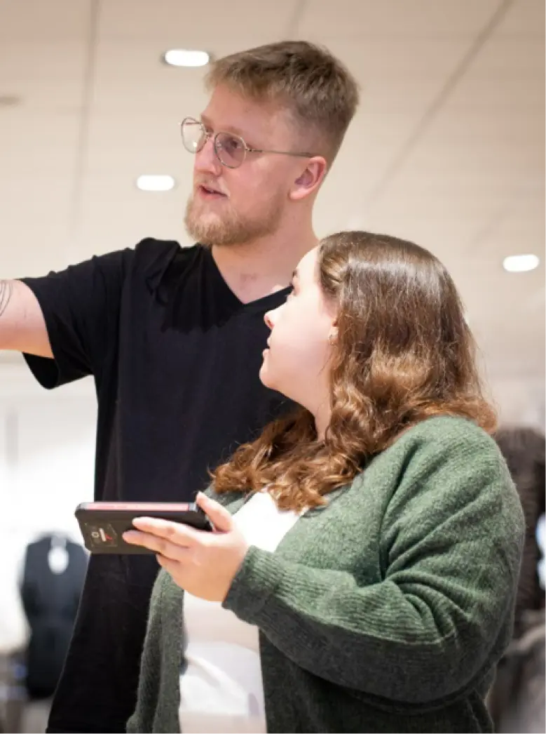 Ein Mann und eine Frau stehen im Haus. Der Mann zeigt geradeaus, während die Frau ein Telefon hält und in die gleiche Richtung blickt. Beide scheinen in ein Gespräch oder eine Zusammenarbeit vertieft zu sein.