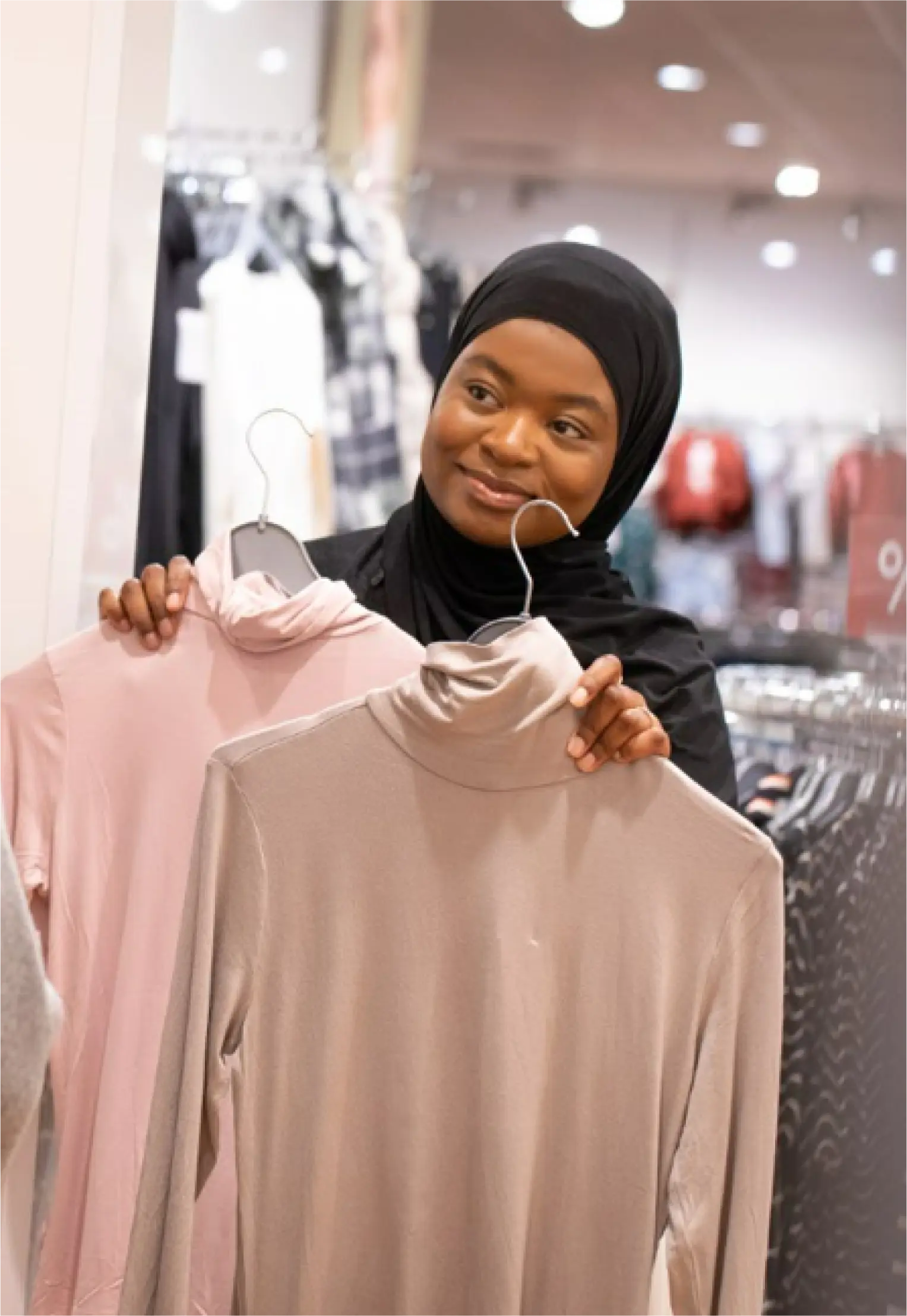 Eine Frau mit schwarzem Hijab lächelt, während sie in einem Bekleidungsgeschäft zwei Rollkragenpullover hochhält, einen rosa und einen beige.