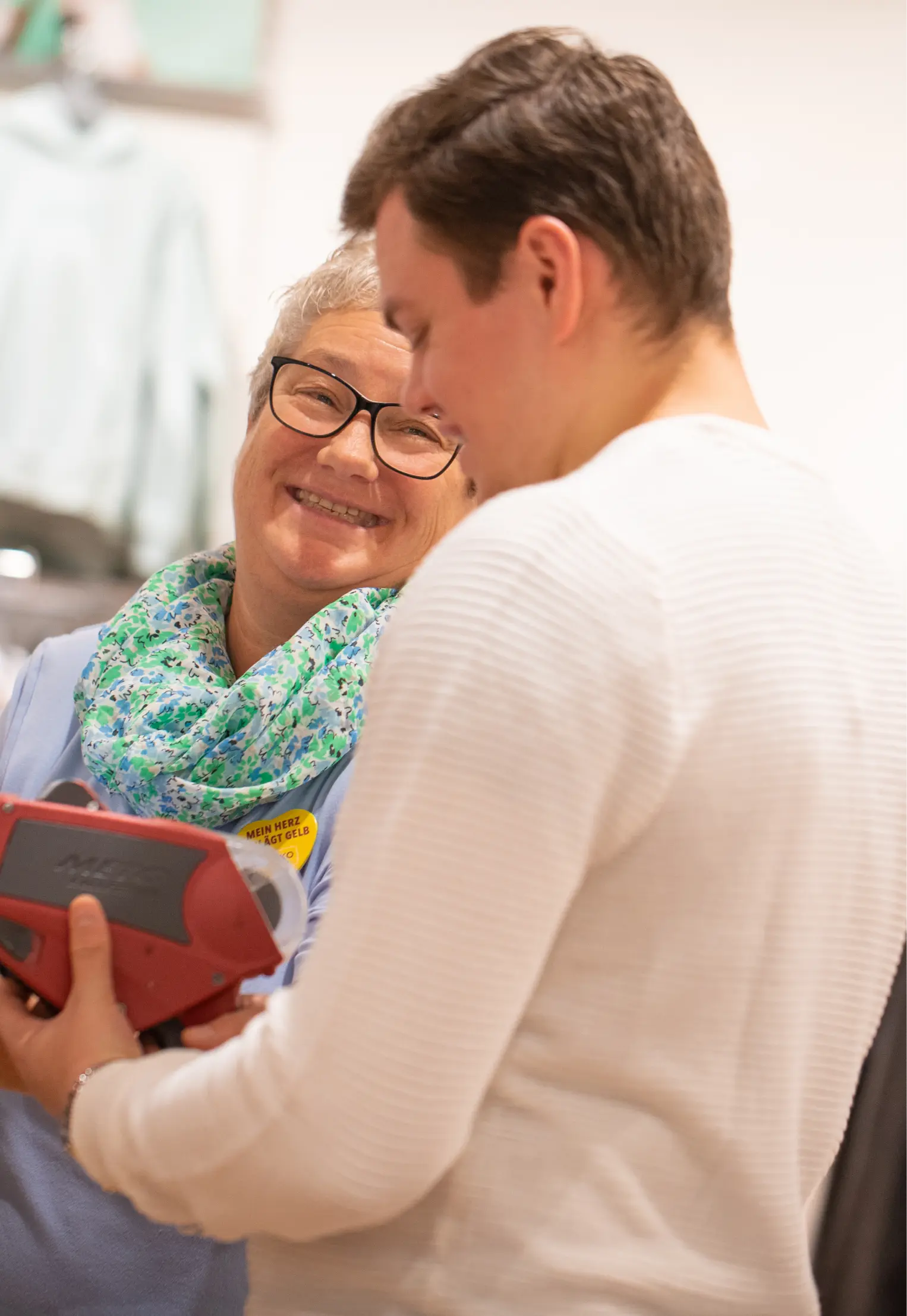 Eine ältere Frau mit Brille und geblümtem Schal lächelt einen Mann im weißen Pullover an, der einen roten Gegenstand in der Hand hält. Sie scheinen sich drinnen nett zu unterhalten.