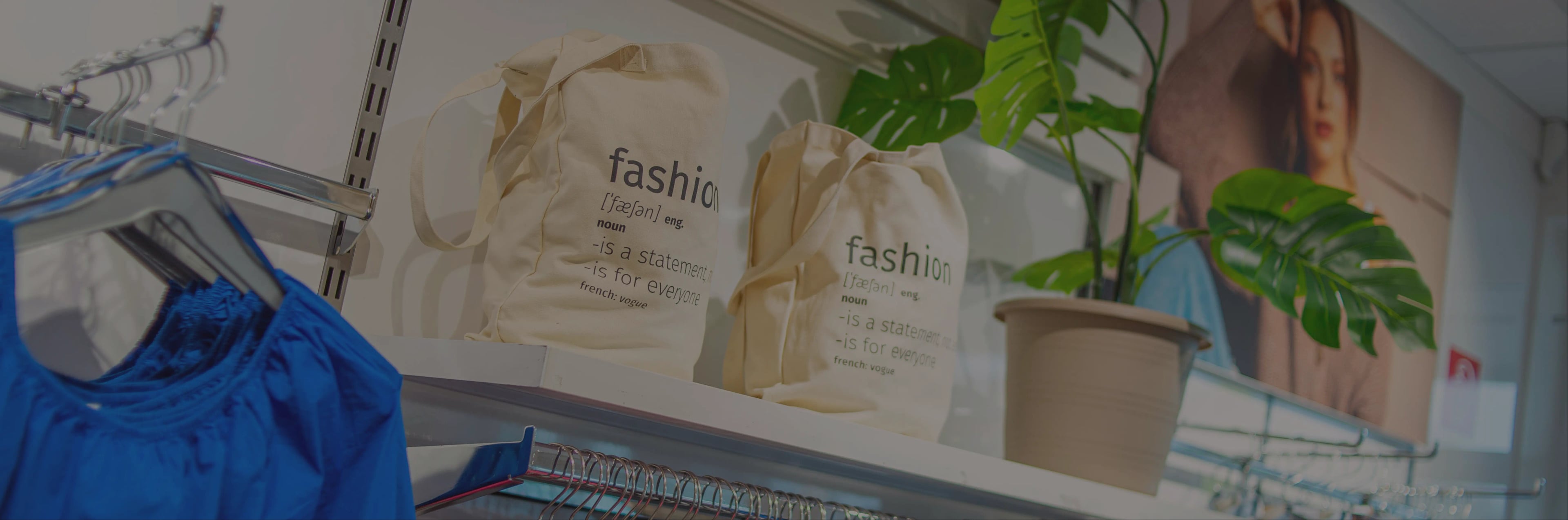 Regaldisplay mit blauer Kleidung, zwei beigen Tragetaschen mit der Aufschrift „Mode“, einer Topfpflanze und einem großen Frauenfoto im Hintergrund.