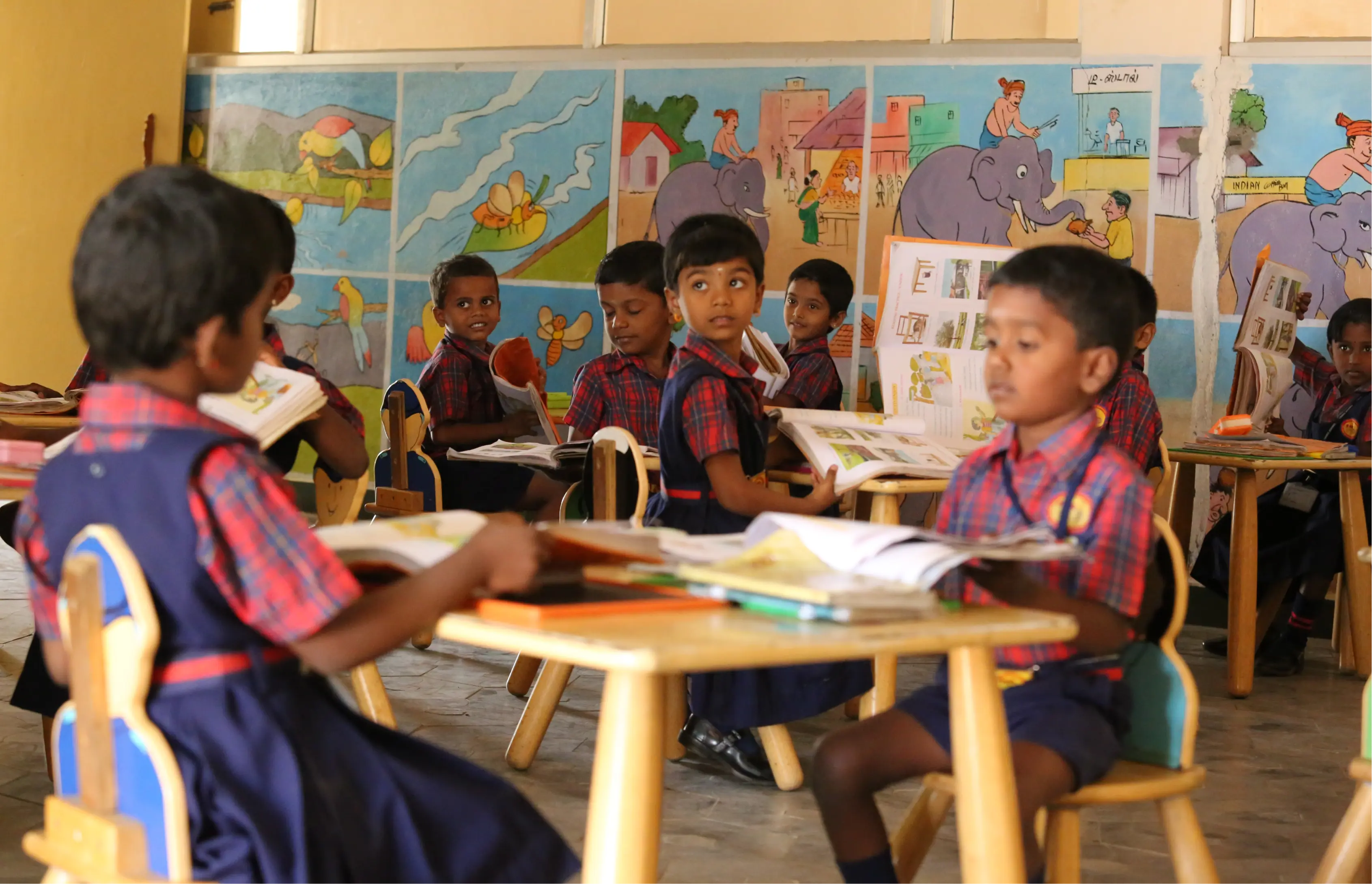Kleine Kinder in rot-blauen Uniformen sitzen in einem farbenfrohen Klassenzimmer an runden Tischen und lesen Bücher. Die Wände sind mit farbenfrohen Gemälden von Tieren und Naturszenen geschmückt.