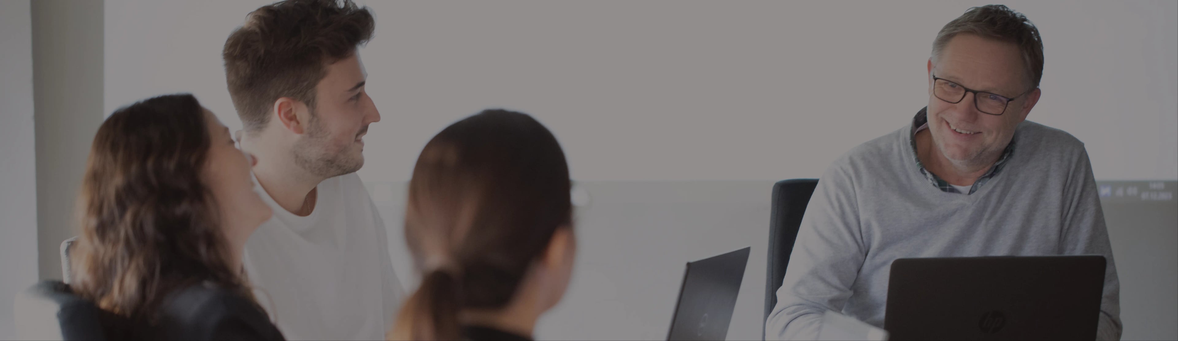 Vier Personen sitzen mit Laptops um einen Tisch und unterhalten sich. Ein Mann mit Brille lächelt, während die anderen ihm aufmerksam zuhören. Die Szene scheint eine Besprechung oder ein gemeinsamer Arbeitsbereich zu sein.