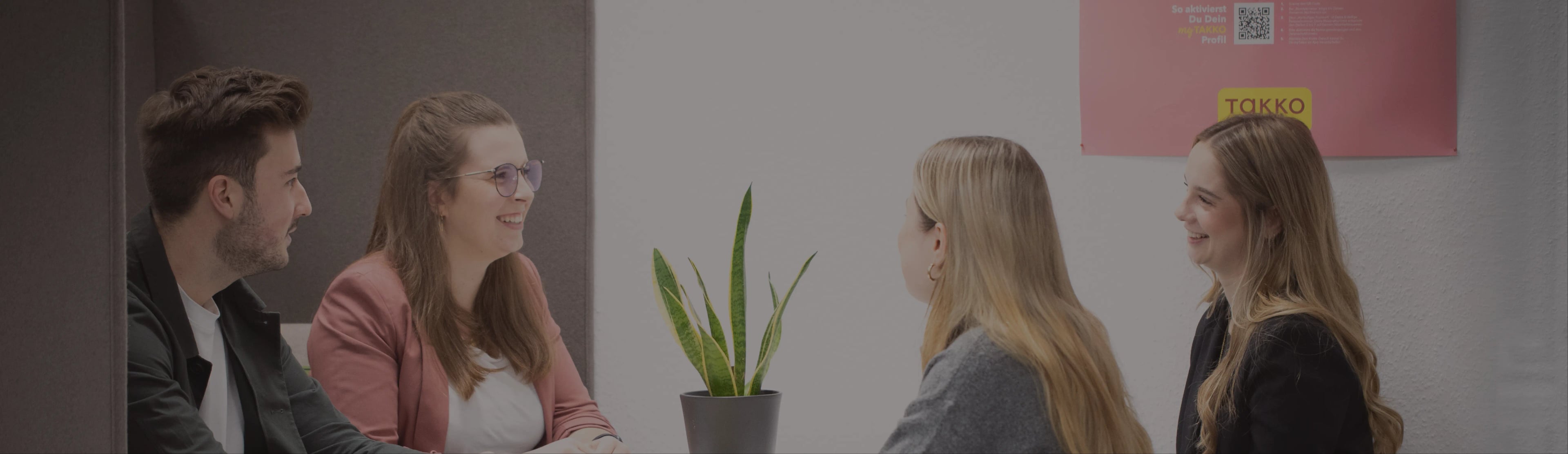 Vier Personen sitzen lächelnd und unterhalten sich um einen Tisch. In der Mitte steht eine Topfpflanze. Im Hintergrund ist an der Wand ein rosa Poster mit Text und einem QR-Code zu sehen.