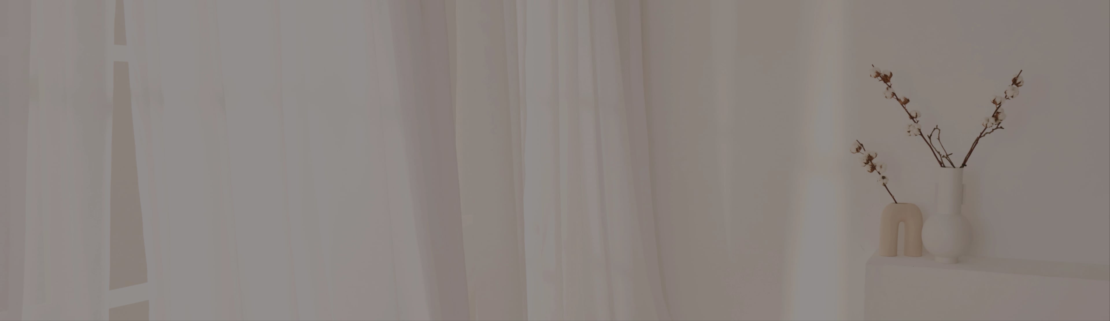 Minimalistisches Interieur mit transparenten weißen Vorhängen und sanfter Beleuchtung. Rechts steht auf einem kleinen weißen Regal eine beige Vase mit zarten Zweigen, an denen kleine weiße und braune Blüten hängen. Das schafft eine ruhige, heitere Atmosphäre.