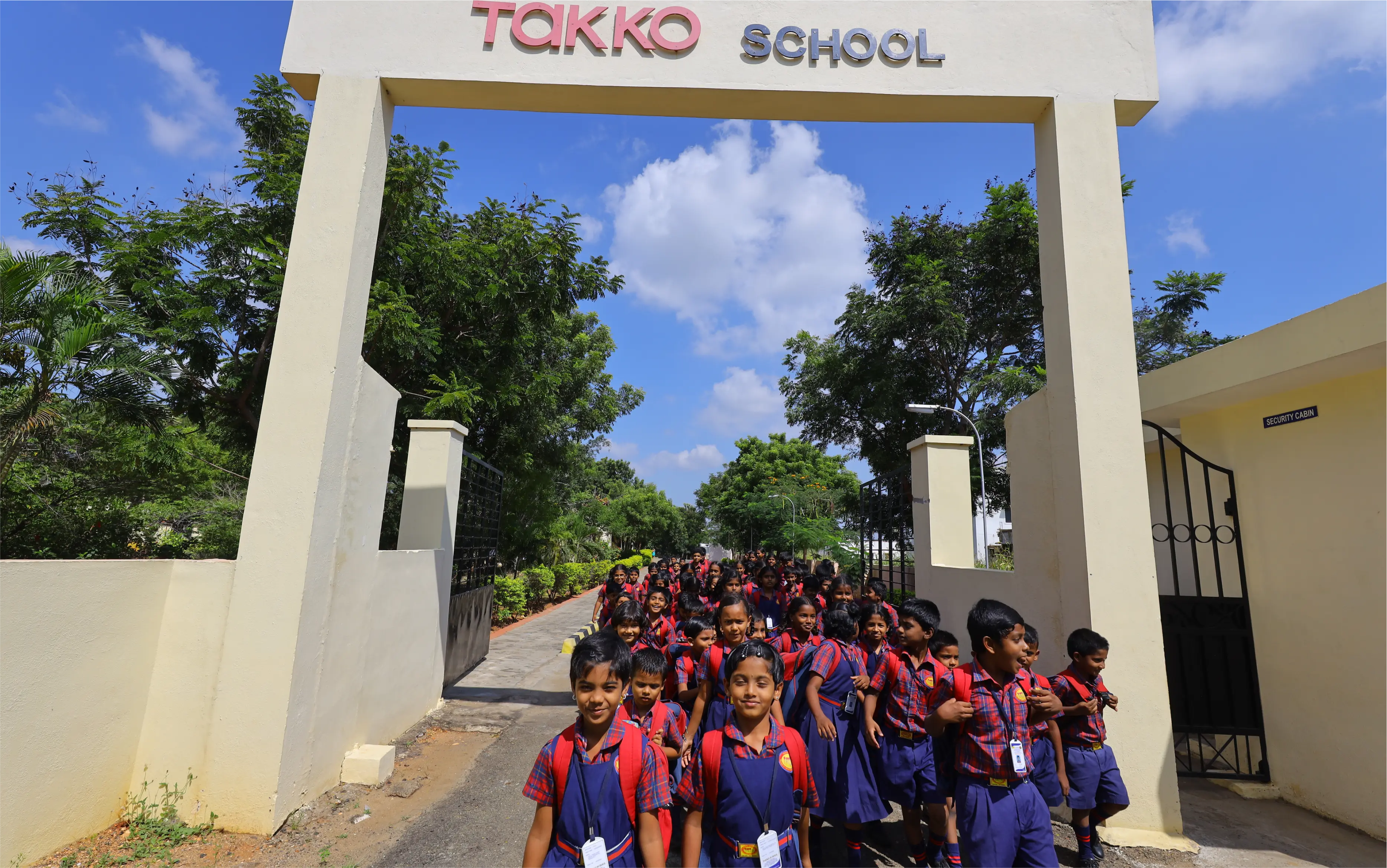 Eine Gruppe junger Schüler in roten und blauen Uniformen kommt an einem sonnigen Tag aus einem Schultor mit der Aufschrift „TAKKO SCHOOL“, umgeben von grünen Bäumen und blauem Himmel.