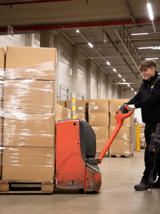 Eine Person bewegt mit einem roten elektrischen Palettenwagen einen großen Stapel in Plastik eingewickelter Kartons in einem geräumigen Lagerhaus.