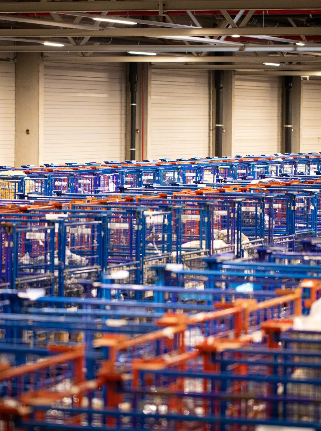Reihen blauer und orangefarbener Metallkarren oder Lagerregale stehen in einer großen Lagerhalle mit weißen Rolltoren im Hintergrund. Die Szene wirkt organisiert und industriell.