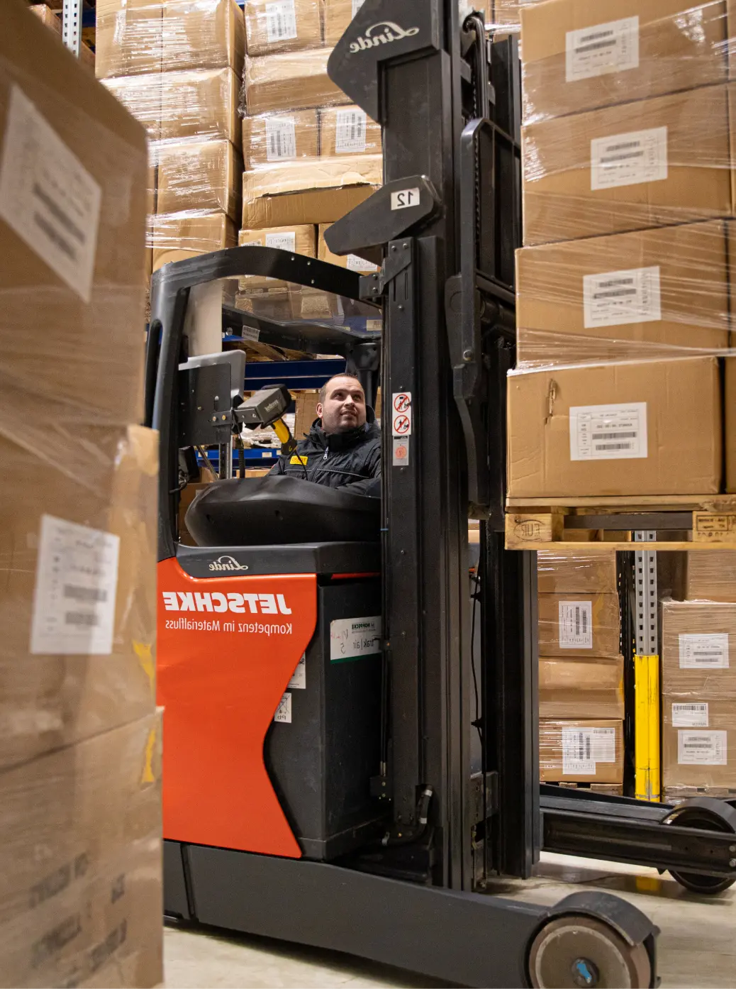 Ein Mann bedient einen Gabelstapler und hebt Kartons in einem Lagerhaus voller gestapelter Pakete und Regale an.
