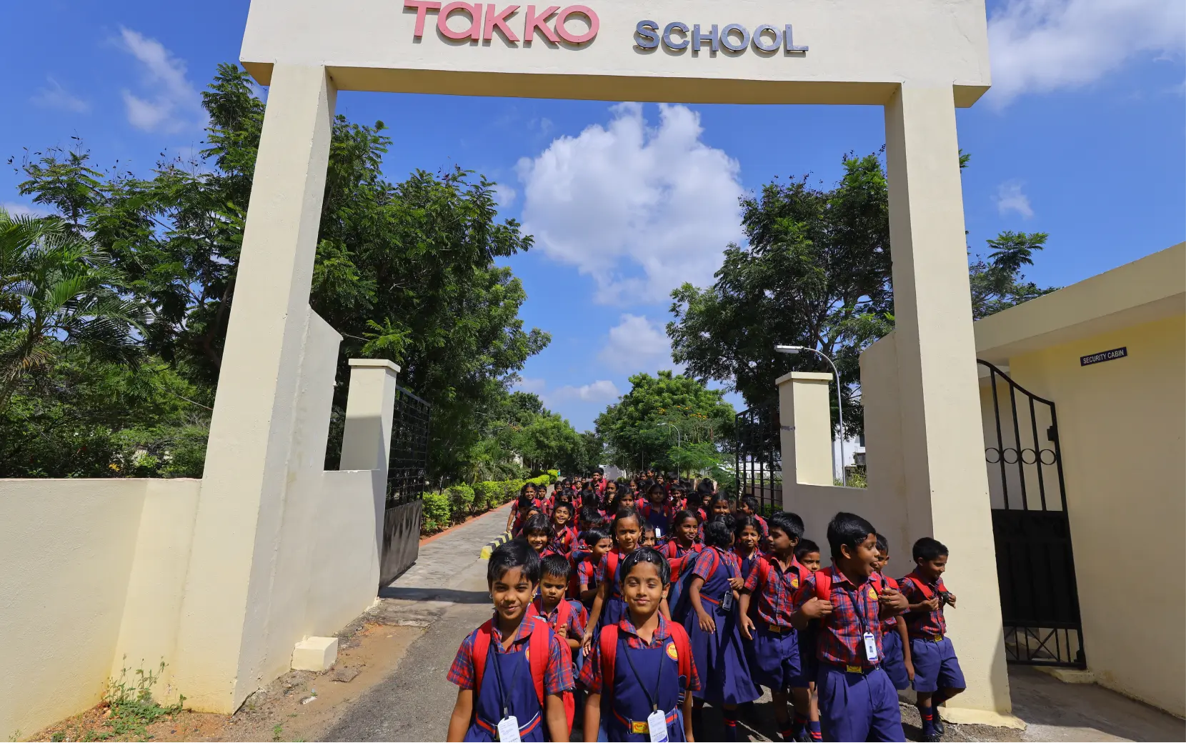 Eine große Gruppe von Kindern in passenden rot-blauen Schuluniformen verlässt an einem sonnigen Tag, umgeben von Bäumen und Grün, das Haupttor einer Schule mit dem Namen „Takko School“.