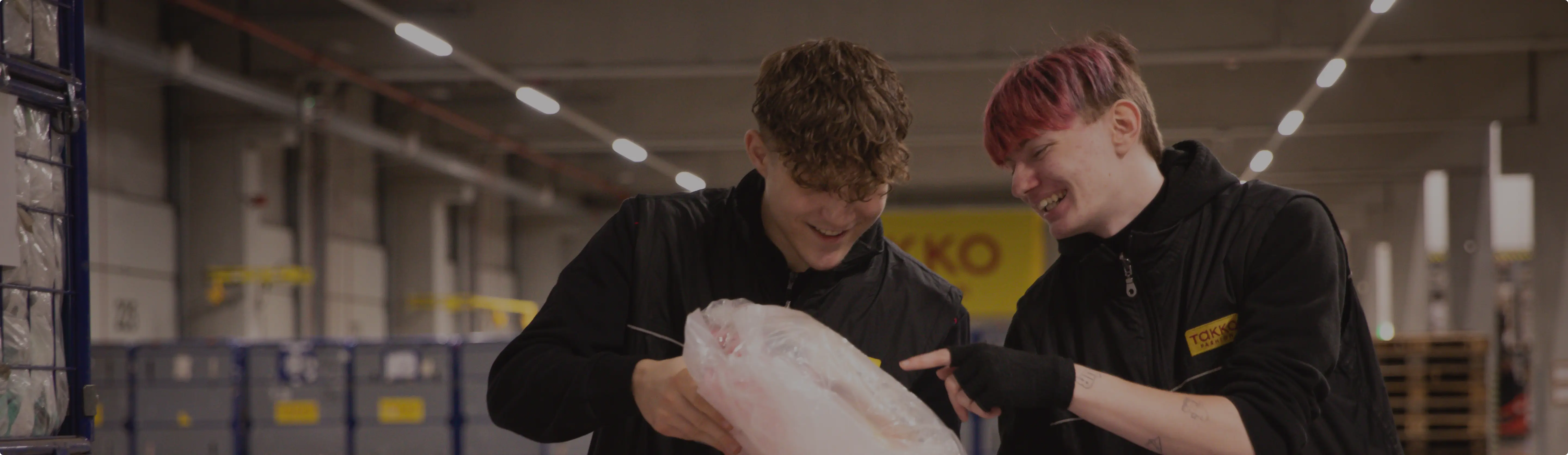 Zwei junge Menschen in schwarzen Uniformen lächeln und untersuchen gemeinsam einen verpackten Artikel in einer Industrielagerhalle mit Lagerregalen und Containern im Hintergrund.