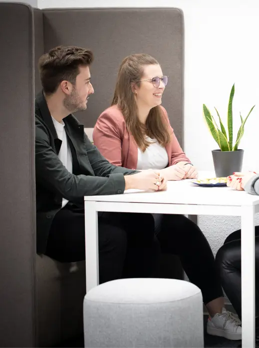 Zwei Personen sitzen nebeneinander in einer Sitznische an einem weißen Tisch, unterhalten sich und lächeln. Eine Topfpflanze steht auf dem Tisch, und ein Teller mit Snacks ist teilweise sichtbar. Die Umgebung erinnert an ein modernes Büro oder einen Besprechungsraum.