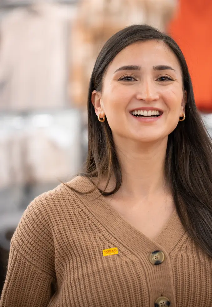 Eine Frau mit langen dunklen Haaren, einer braunen Strickjacke und kleinen Creolen lächelt in die Kamera. An ihrer Strickjacke trägt sie ein gelbes Namensschild. Der Hintergrund ist leicht verschwommen.