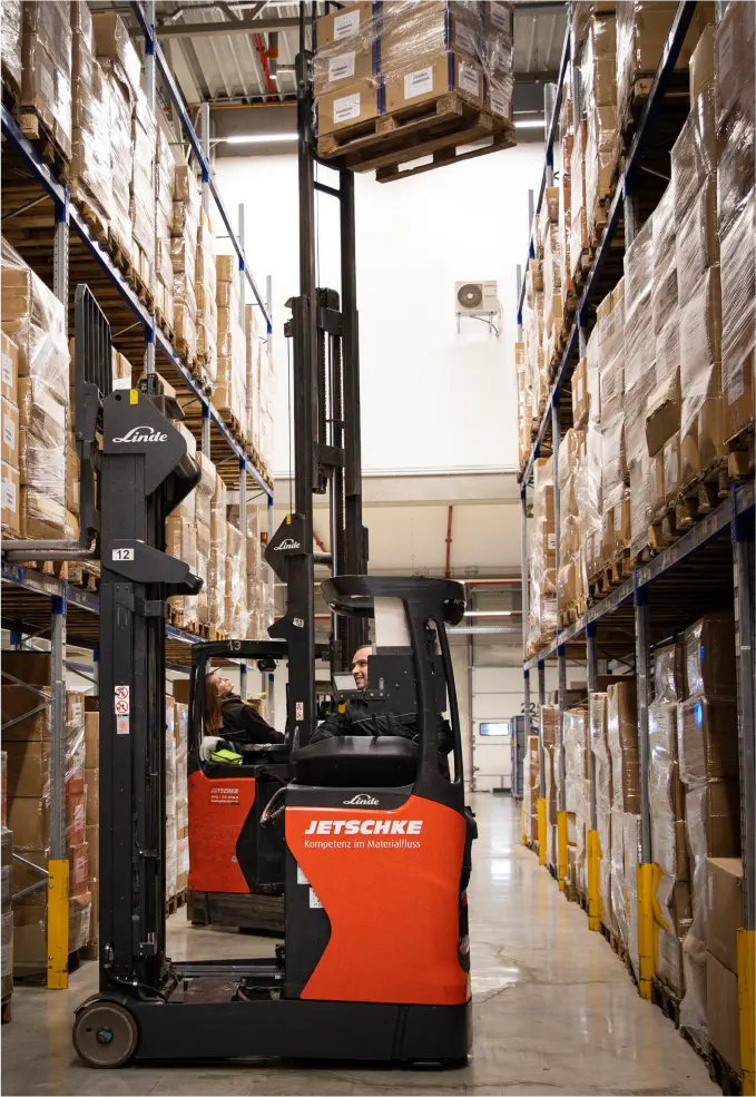 Ein Arbeiter bedient einen roten Gabelstapler, um eine Palette mit Kartons auf ein Regal in einem Lagergang zu heben, der mit hohen Regalen voller brauner Kartons gesäumt ist.