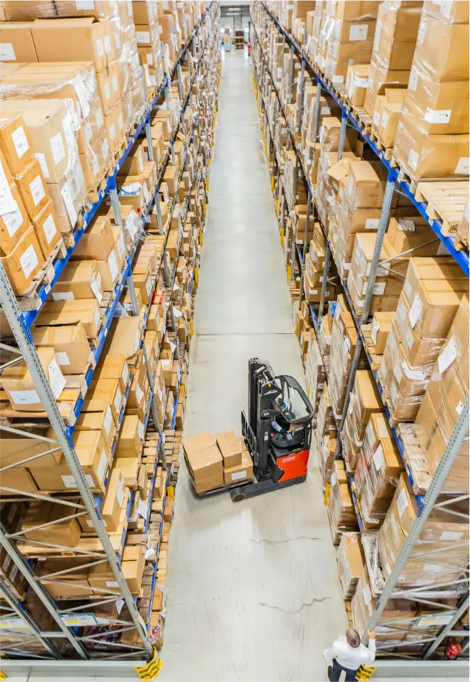 Ein Gabelstaplerfahrer bewegt Kisten in einem schmalen Gang zwischen hohen, mit Kartons gefüllten Regalen in einem großen Lagerhaus (von oben gesehen).