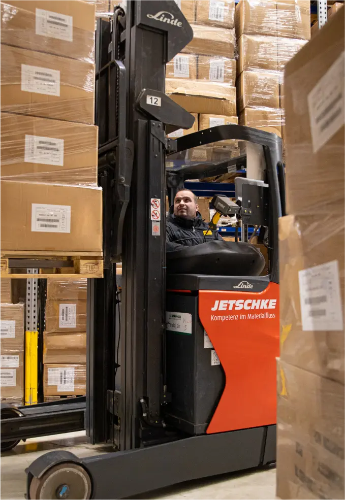 Ein Mann bedient einen Gabelstapler in einem Lagerhaus und manövriert zwischen hohen Stapeln von Kartons in Regalen. Der Gabelstapler trägt die Marken Linde und Jetschke und ist von ordentlich sortierten Paketen umgeben.