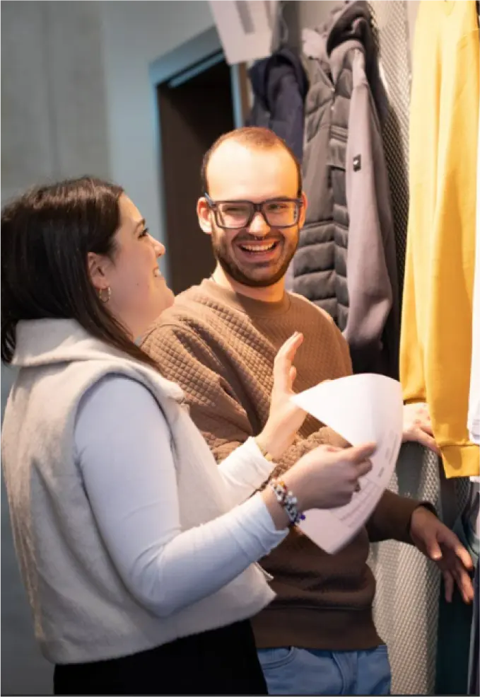 Zwei Personen stehen im Raum, lächeln und unterhalten sich. Einer hält Papiere in der Hand, der andere trägt eine Brille und einen braunen Pullover. Mäntel und Jacken hängen daneben an der Wand. Die Atmosphäre wirkt freundlich und locker.