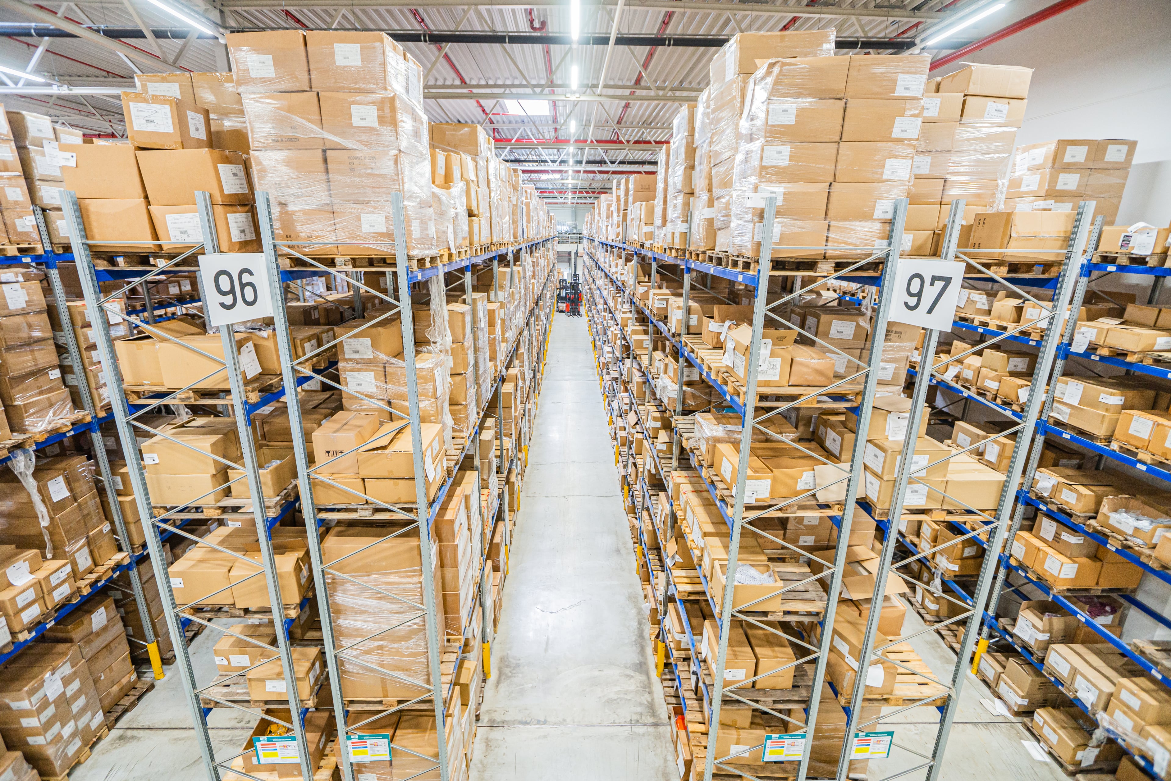Weitwinkelansicht eines Lagerhalleninneren mit hohen Regalen voller Kartons zu beiden Seiten eines Mittelgangs. Schilder mit den Aufschriften 96 und 97 hängen an den Regalen. Helle Deckenbeleuchtung erhellt den Raum.