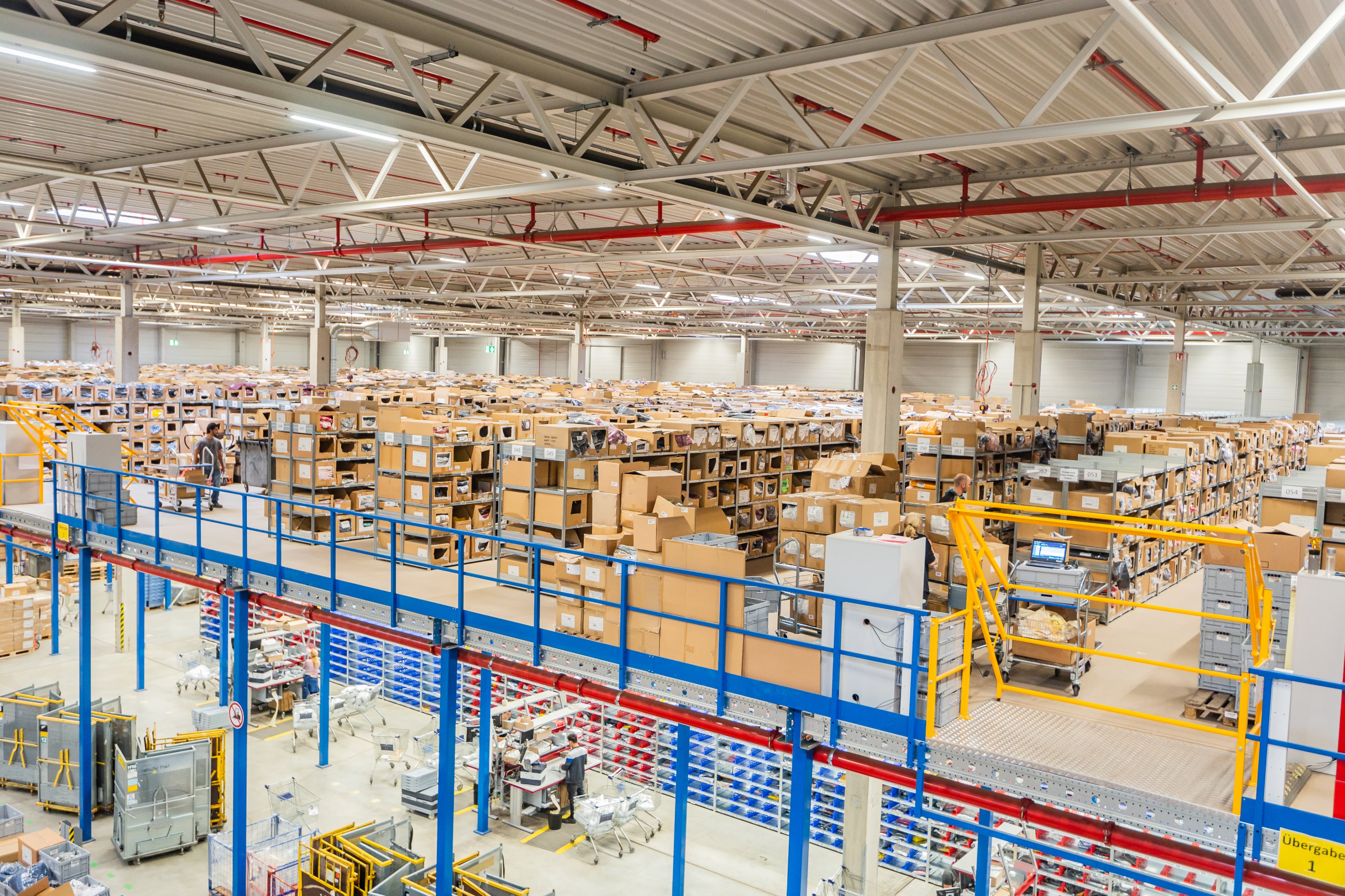 Der Innenraum einer großen Lagerhalle mit hohen Regalen voller ordentlich gestapelter Kisten und Pakete, sichtbarer Industriebeleuchtung und einem Laufsteg über den Lagerbereichen.
