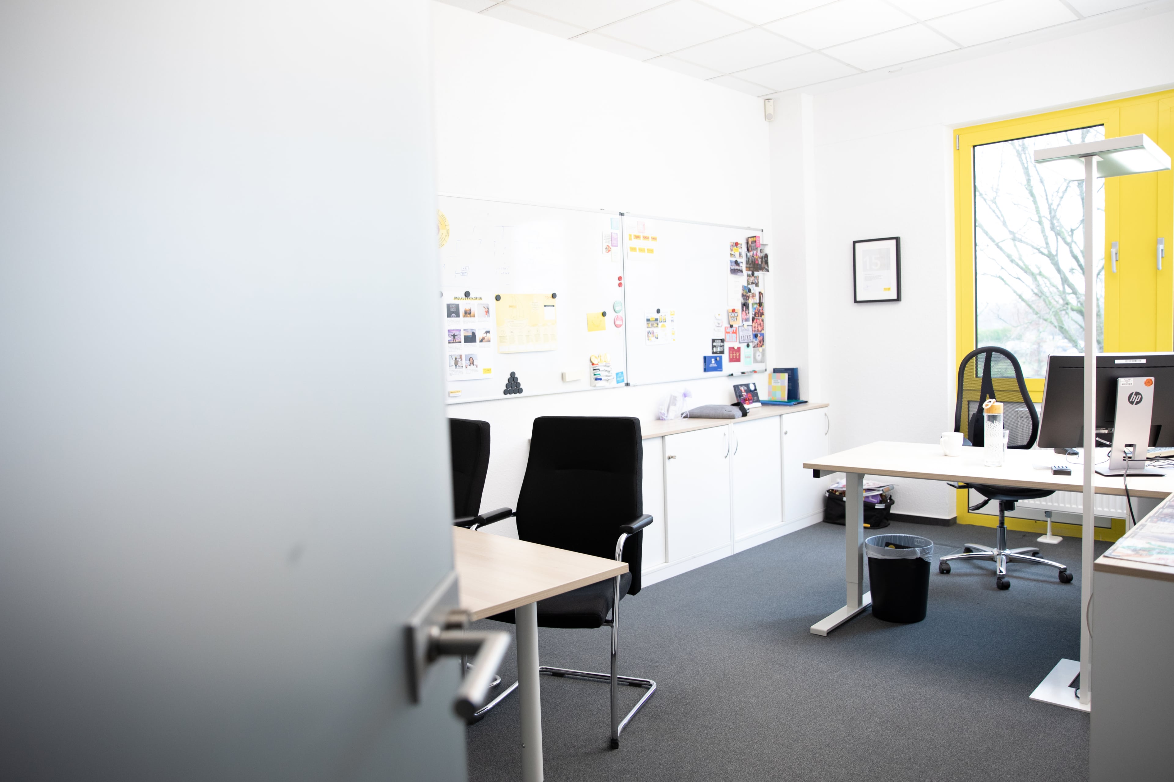 Ein helles, modernes Büro mit zwei Schreibtischen, Bürostühlen, einem Computermonitor, einem Whiteboard mit Notizen und Bildern und einem großen Fenster mit gelbem Rahmen, das natürliches Licht hereinlässt.
