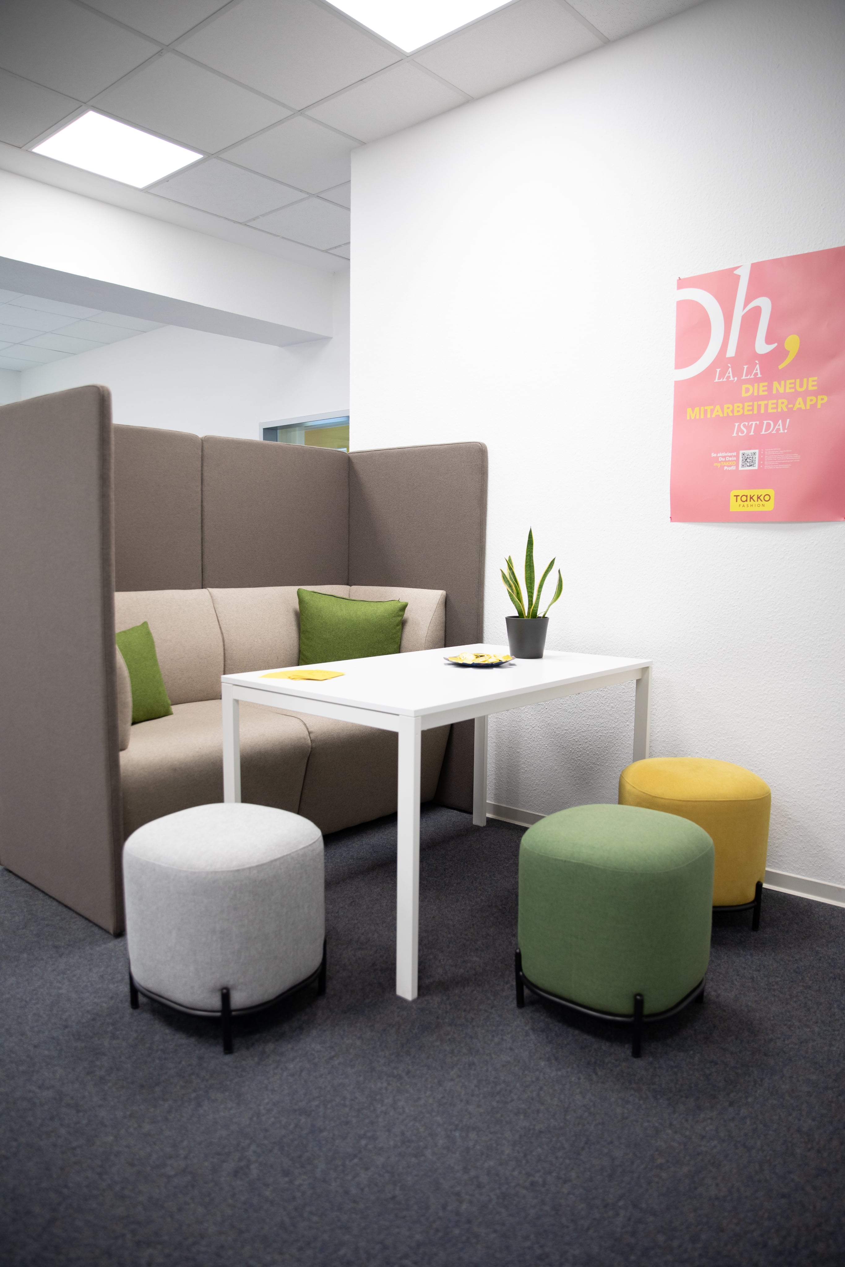 Ein moderner Bürositzbereich mit einer gepolsterten Sitzecke, einem weißen Tisch und drei runden Hockern in Grau, Grün und Gelb. Auf der Sitzecke liegen grüne Kissen, an der Wand hängt ein rosa Poster.