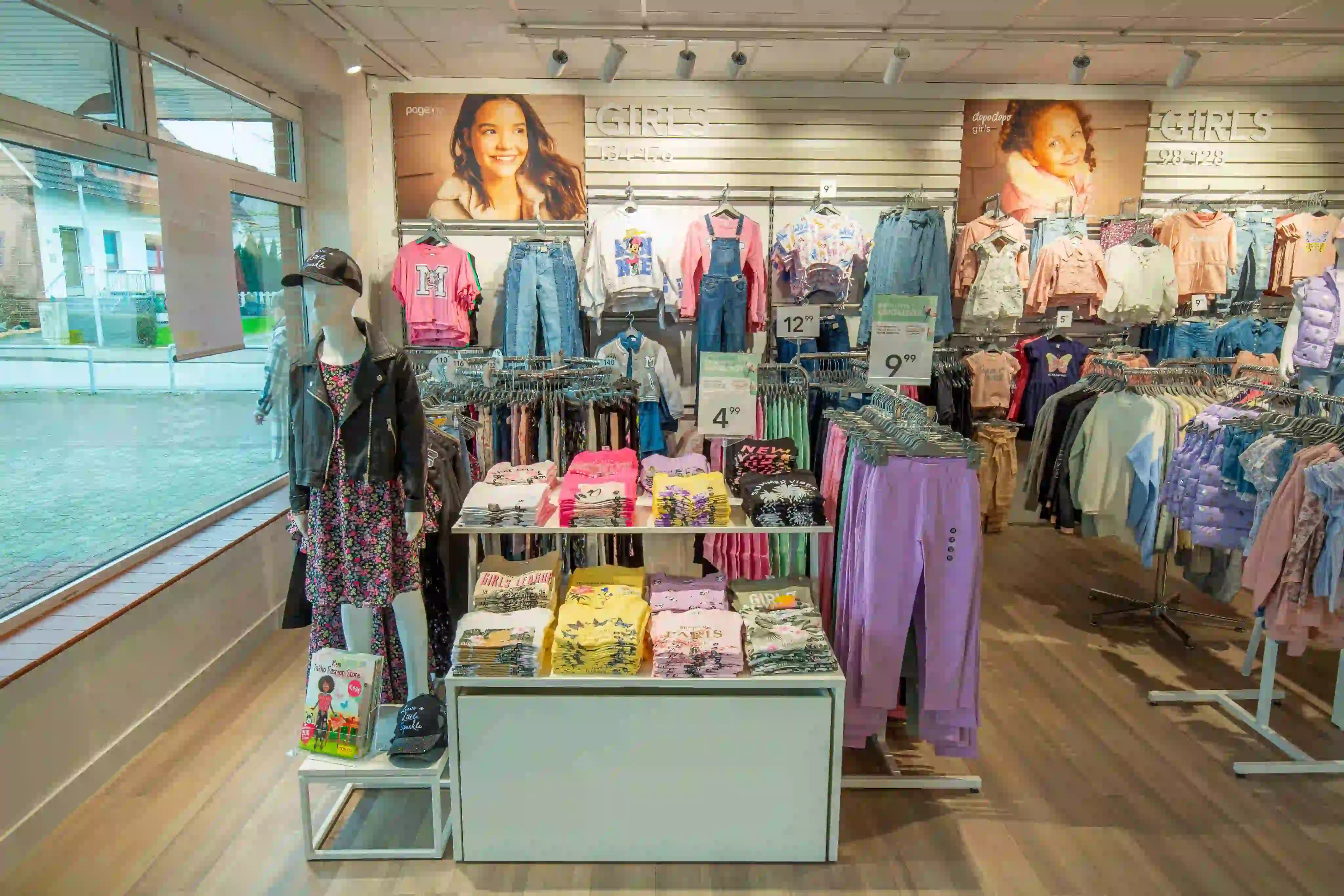 Kinderbekleidungsabteilung mit Regalen voller bunter Hemden, Hosen und Jacken. Eine Schaufensterpuppe trägt ein Kleid und eine Lederjacke. An der Wand hängen Poster von Mädchen, die Outfits vorführen. Große Fenster lassen Tageslicht herein.