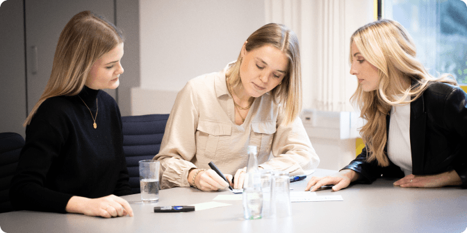 Drei Frauen sitzen an einem Tisch in einem Büro und konzentrieren sich auf Papierkram. Eine Frau schreibt, während die anderen beiden zuschauen. Auf dem Tisch stehen Gläser und eine Flasche Wasser. Die Atmosphäre wirkt professionell und kooperativ.
