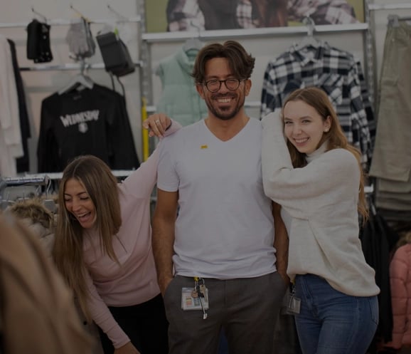 Drei Menschen stehen lächelnd in einem Bekleidungsgeschäft. Der Mann in der Mitte trägt eine Brille und ein weißes Hemd, während die beiden leger gekleideten Frauen neben ihm sich zu ihm beugen und fröhlich und glücklich wirken. Hinter ihnen sind Kleiderständer zu sehen.