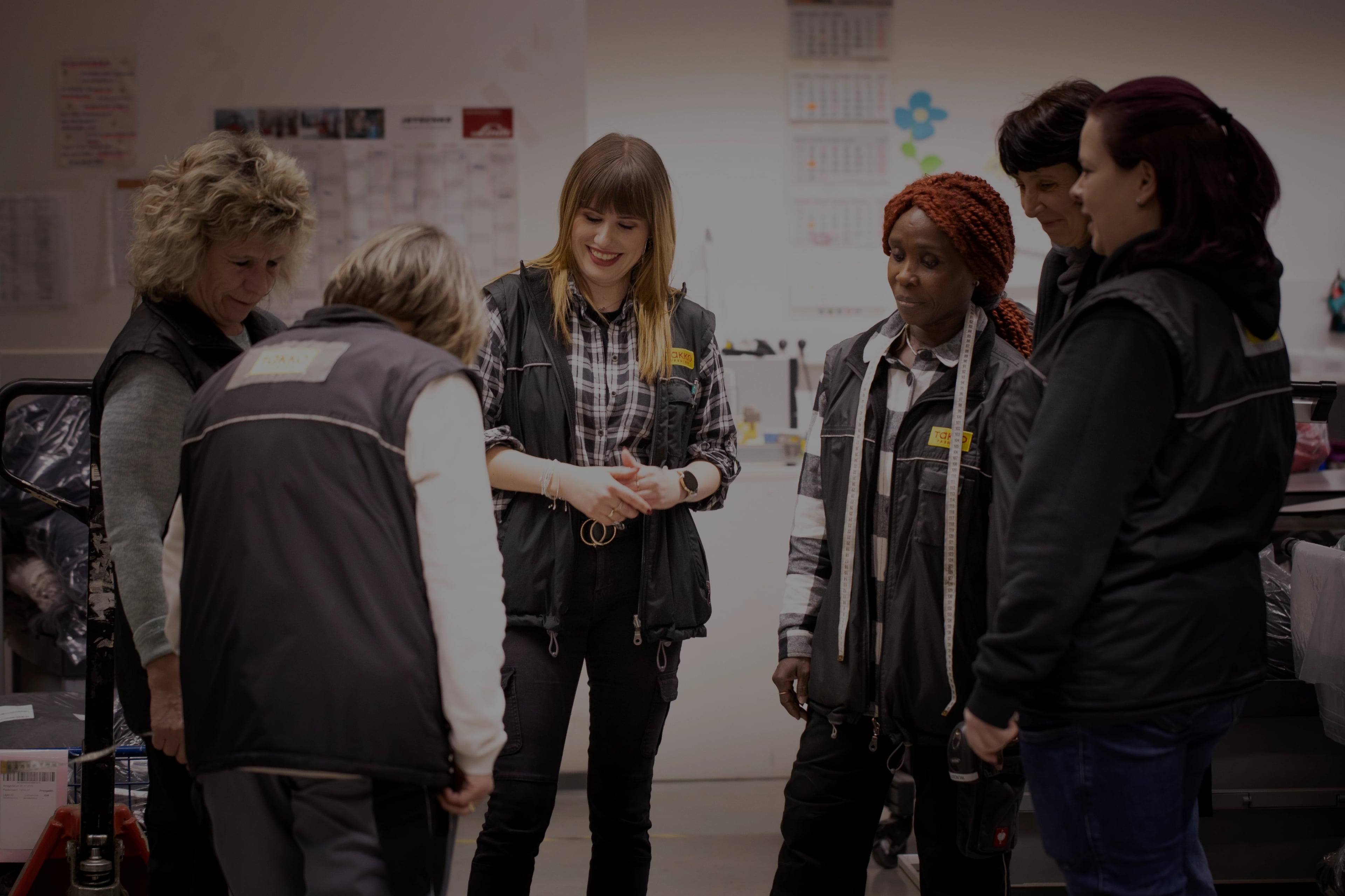 Sechs Frauen in dunklen Westen stehen in einem hell erleuchteten Raum zusammen und scheinen ein freundliches Gespräch zu führen. Einige lächeln, und eine beugt sich leicht zur Mitte der Gruppe.