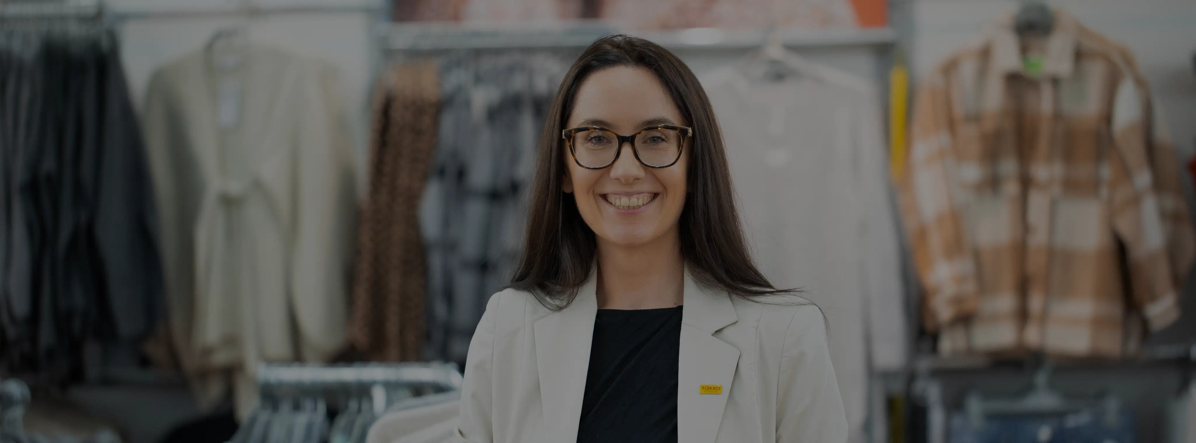 Eine Frau mit langen braunen Haaren und Brille, die einen weißen Blazer und ein schwarzes Hemd trägt, lächelt, während sie in einem Bekleidungsgeschäft steht, hinter dem sich Kleiderständer mit Kleidung befinden.