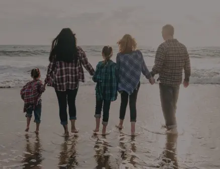 Fünf Menschen – zwei Erwachsene und drei Kinder – gehen Hand in Hand an einem Strand entlang, mit Blick aufs Meer. Sie tragen legere, karierte Hemden und Jeans, und ihre Spiegelbilder sind im nassen Sand sichtbar.