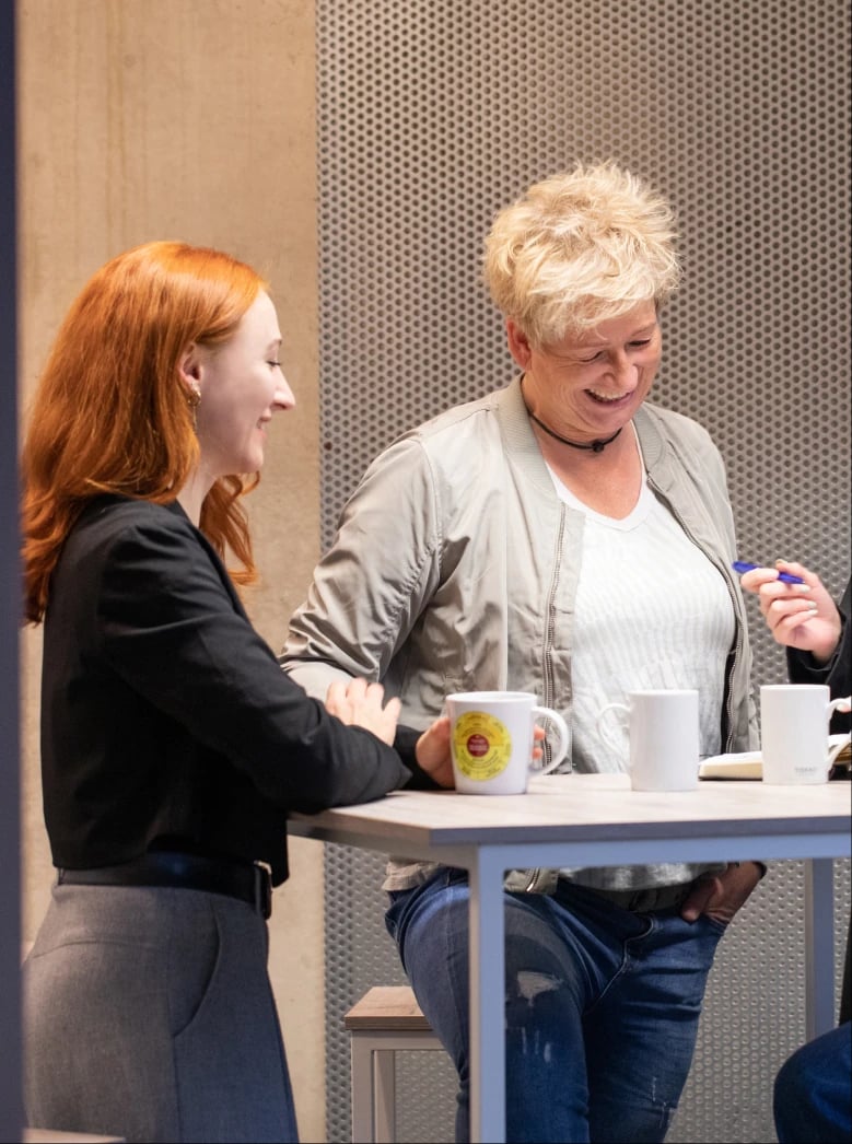 Zwei Frauen stehen lächelnd und unterhalten sich an einem hohen Tisch. Eine hält einen Stift über einem Notizbuch, auf dem Tisch stehen zwei Kaffeetassen. Im Hintergrund ist eine graue Lochwand zu sehen.