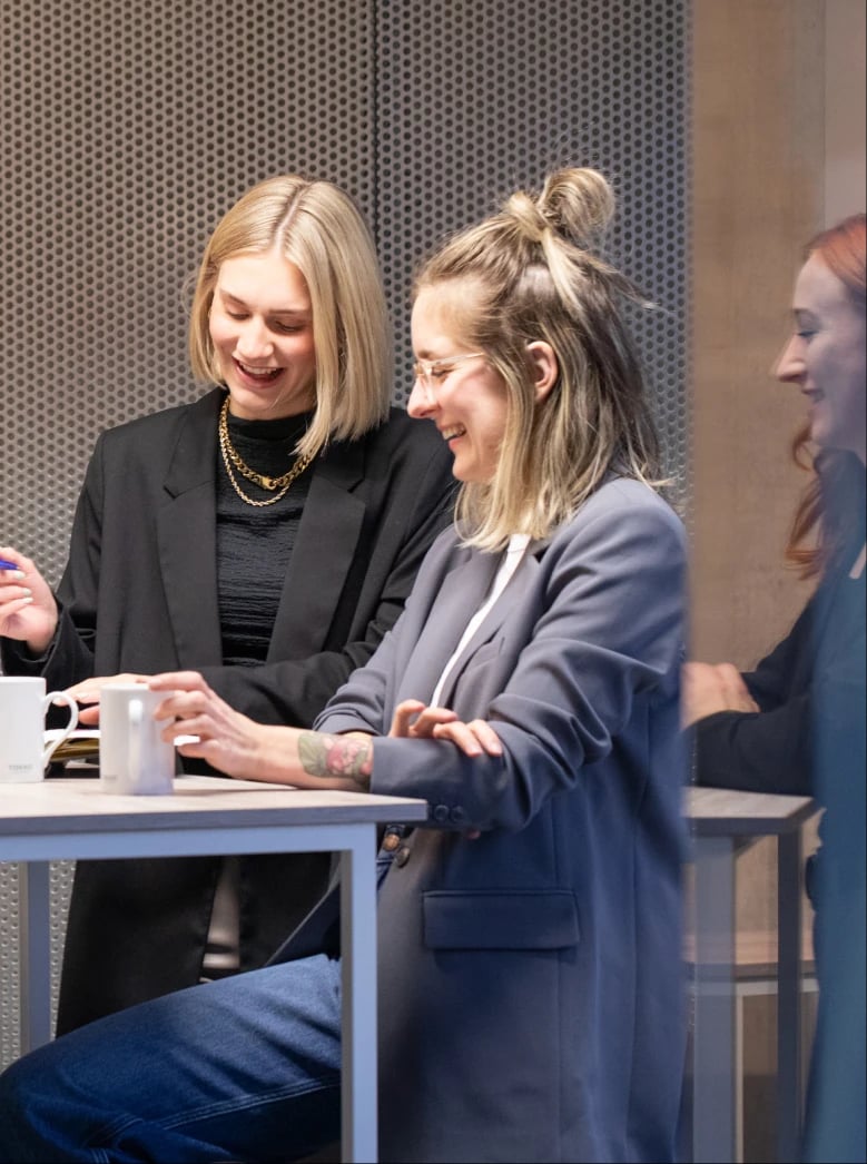 Zwei Frauen sitzen lächelnd und unterhalten sich an einem Tisch. Die eine hält einen Stift, die andere eine Tasse. Eine dritte Frau ist im Hintergrund leicht verschwommen zu sehen. Sie scheinen sich in einem modernen Büro zu befinden.