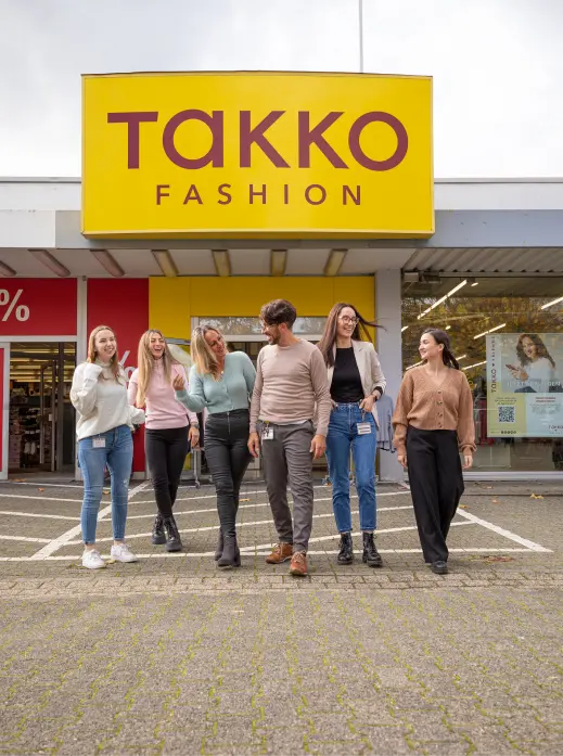 Sechs Menschen stehen und gehen lächelnd vor einem Takko Fashion-Geschäft mit einem großen gelb-roten Schild über dem Eingang.